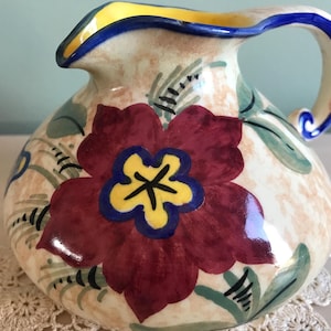 May include: A ceramic pitcher with a floral design. The pitcher is cream colored with a red flower with a yellow center and blue accents. The pitcher has a blue and yellow rim and a blue handle.