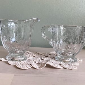 May include: A clear glass creamer and sugar bowl set with a delicate floral pattern. The creamer has a single handle and the sugar bowl has two handles.