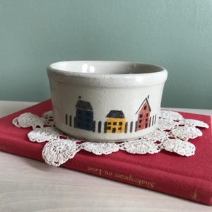 May include: A small, off-white ceramic bowl with a hand-painted village scene featuring blue, yellow, and red houses. The bowl is placed on a red book with a white lace doily. The bowl is approximately 4 inches in diameter.