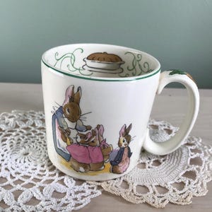 May include: White ceramic mug featuring a charming illustration of rabbits in a storybook scene. The mug has a green rim and handle with a carrot design. The artwork depicts a pie and a family of rabbits, perfect for a cozy beverage.