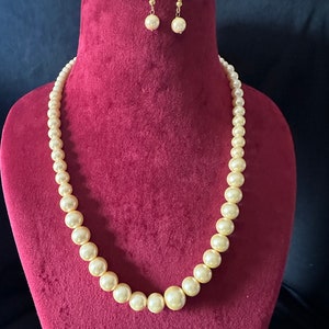 May include: A cream pearl necklace and matching earrings set. The necklace has graduated pearls, with larger pearls at the centre and smaller ones towards the clasp. The earrings have a single pearl suspended from gold-coloured hooks. The jewellery is displayed on a burgundy velvet bust.