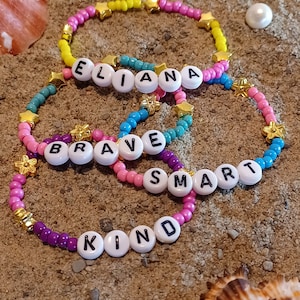 May include: Four colorful beaded bracelets with gold stars and white letter beads spelling out the words "Eliana", "Brave", "Smart", and "Kind".