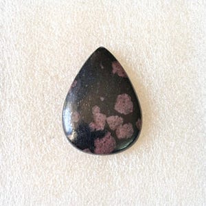 Puede incluir: Una gema en forma de l&aacute;grima con superficie pulida. La piedra es principalmente negra con grupos de inclusiones de color rojo p&uacute;rpura. La gema est&aacute; sobre un fondo blanco.