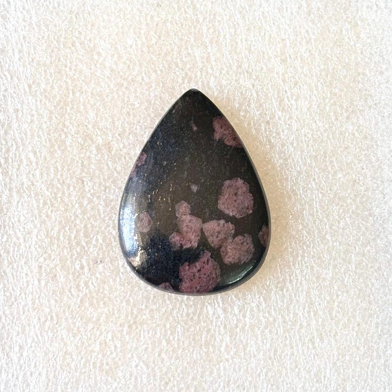 Puede incluir: Una gema en forma de l&aacute;grima con superficie pulida. La piedra es principalmente negra con grupos de inclusiones de color rojo p&uacute;rpura. La gema est&aacute; sobre un fondo blanco.