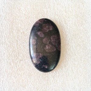 May include: An oval-shaped, polished gemstone with a dark, almost black base color. The stone is speckled with clusters of deep purple or burgundy-colored inclusions, creating a striking contrast. The surface is smooth and reflective.