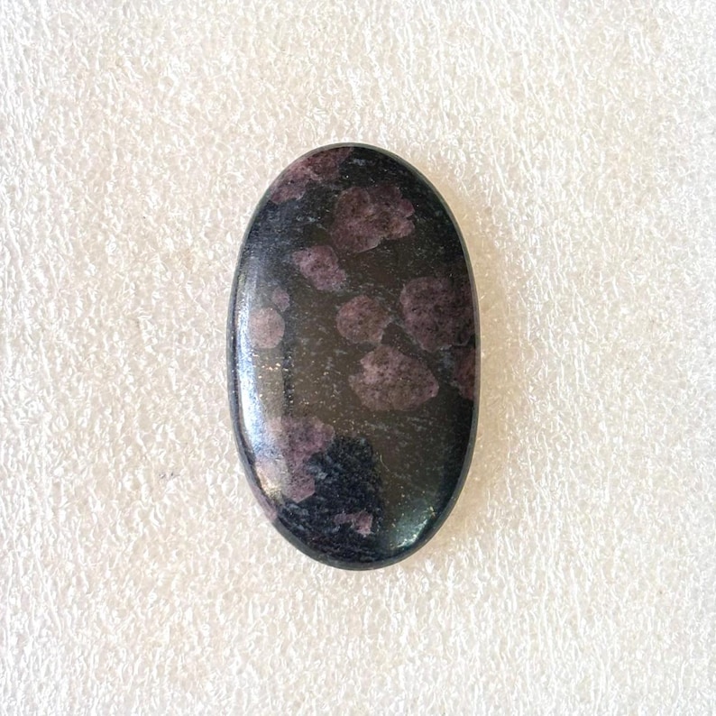 May include: An oval-shaped, polished gemstone with a dark, almost black base color. The stone is speckled with clusters of deep purple or burgundy-colored inclusions, creating a striking contrast. The surface is smooth and reflective.