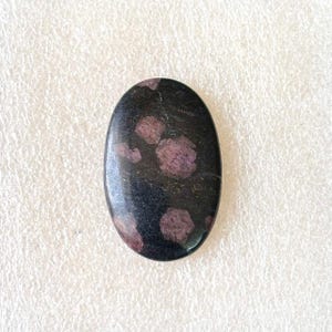 Puede incluir: Una piedra preciosa ovalada con una superficie pulida. La piedra es principalmente negra con grupos de manchas de color rojo p&uacute;rpura. La piedra preciosa est&aacute; sobre un fondo blanco.