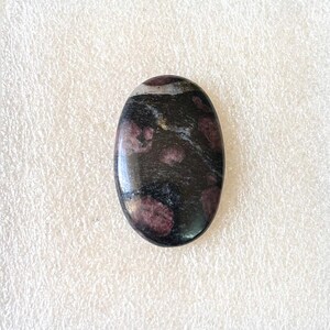 May include: An oval-shaped gemstone with a polished surface. The stone features a dark base color with maroon and white speckled patterns. The gemstone is likely used for jewelry or decorative purposes.