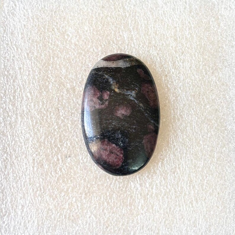 May include: An oval-shaped gemstone with a polished surface. The stone features a dark base color with maroon and white speckled patterns. The gemstone is likely used for jewelry or decorative purposes.