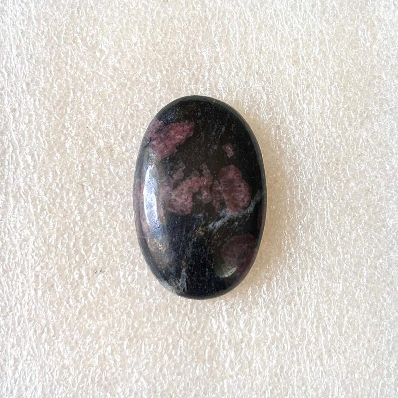 Puede incluir: Gema de forma ovalada con superficie pulida. La piedra es principalmente negra con parches irregulares de rojo intenso y toques de blanco. La gema probablemente se utiliza para joyer&iacute;a o fines decorativos.