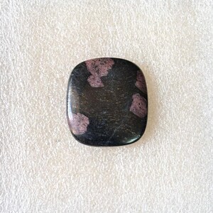 Puede incluir: Una gema cuadrada pulida con esquinas redondeadas. Predomina el color negro con parches irregulares de rosa intenso o borgo&ntilde;a. La superficie es lisa y reflectante, lo que sugiere un acabado de alta calidad. Ideal para joyer&iacute;a.
