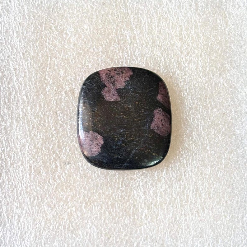Puede incluir: Una gema cuadrada pulida con esquinas redondeadas. Predomina el color negro con parches irregulares de rosa intenso o borgo&ntilde;a. La superficie es lisa y reflectante, lo que sugiere un acabado de alta calidad. Ideal para joyer&iacute;a.