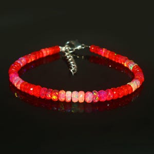Puede incluir: Un brazalete de piedras preciosas de ópalo rojo y rosa con un cierre plateado. El brazalete está hecho de pequeñas cuentas redondas y tiene un degradado de color de rojo a rosa.