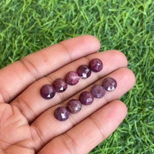 May include: A collection of faceted, round, dark red gemstones. The gems are arranged in rows on a hand, with a green grassy background. The gems have a polished, reflective surface, and the word "Gemswan" is visible.