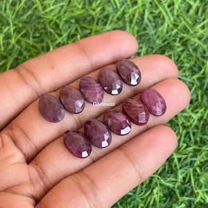 Op de afbeelding: Een verzameling ovaalvormige, gefacetteerde edelstenen in verschillende tinten diep rood en paars. De edelstenen worden in een hand gehouden tegen een achtergrond van groen gras. De edelstenen hebben een gepolijst, reflecterend oppervlak.