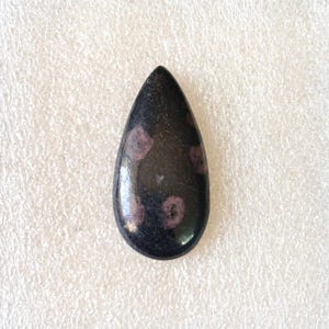 Puede incluir: Una gema en forma de l&aacute;grima pulida y brillante. La piedra es principalmente negra con varias peque&ntilde;as inclusiones redondas de color p&uacute;rpura rojizo. La gema es lisa y parece ser un cabuj&oacute;n.