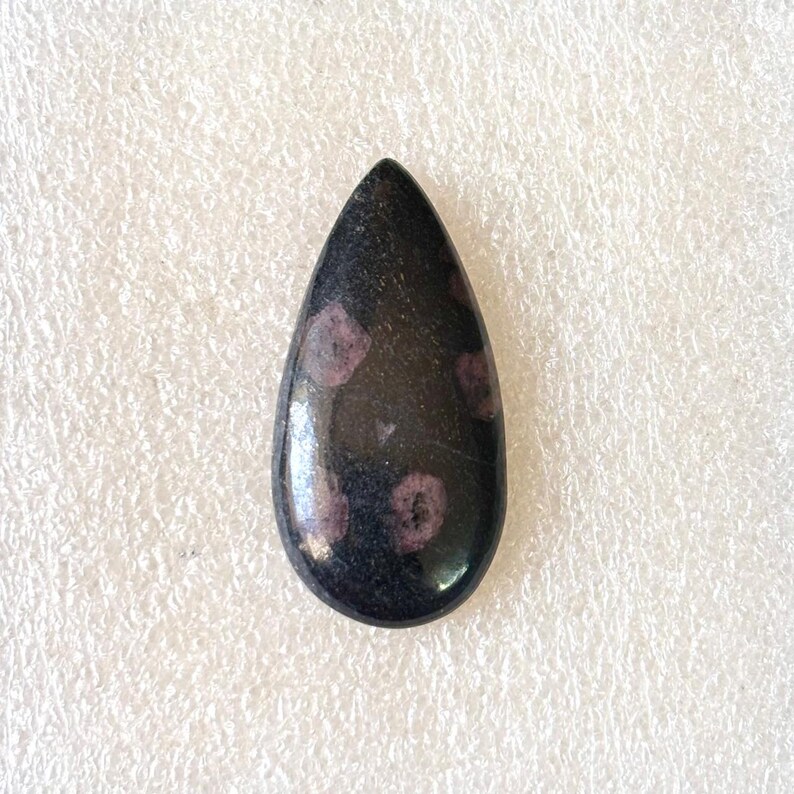 Puede incluir: Una gema en forma de l&aacute;grima pulida y brillante. La piedra es principalmente negra con varias peque&ntilde;as inclusiones redondas de color p&uacute;rpura rojizo. La gema es lisa y parece ser un cabuj&oacute;n.