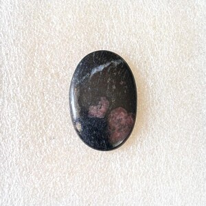 Puede incluir: Gema ovalada con superficie pulida. La piedra es principalmente negra con parches de rojo intenso y vetas blancas. El cabuj&oacute;n liso es adecuado para la creaci&oacute;n de joyas o para coleccionar.