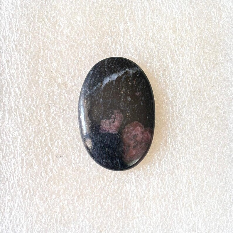 Puede incluir: Gema ovalada con superficie pulida. La piedra es principalmente negra con parches de rojo intenso y vetas blancas. El cabuj&oacute;n liso es adecuado para la creaci&oacute;n de joyas o para coleccionar.