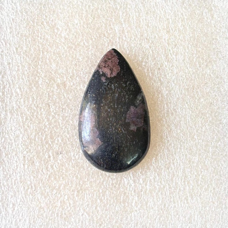 Puede incluir: Una gema pulida en forma de l&aacute;grima. La piedra es predominantemente negra, con &aacute;reas de color rosa intenso y p&uacute;rpura. Se encuentra sobre un fondo blanco.