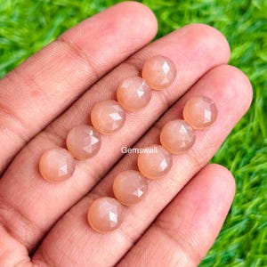 May include: A collection of peach-colored, faceted gemstones, possibly moonstones, are displayed in a hand against a green background. The gemstones are round and have a polished, reflective surface. The text "Gemswali" is visible.
