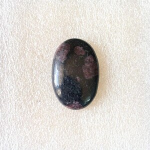 Puede incluir: Gema ovalada con superficie pulida. La piedra es principalmente negra con inclusiones de color rojo oscuro y p&uacute;rpura. La gema est&aacute; sobre un fondo blanco.