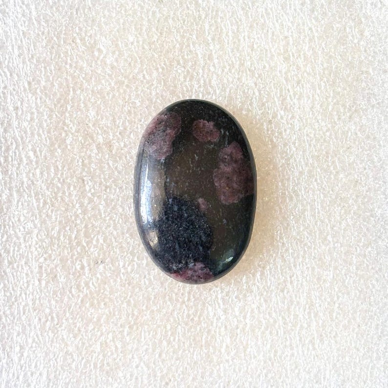 Puede incluir: Gema ovalada con superficie pulida. La piedra es principalmente negra con inclusiones de color rojo oscuro y p&uacute;rpura. La gema est&aacute; sobre un fondo blanco.