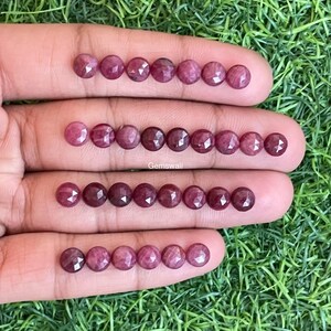 May include: Rows of faceted, round, ruby-colored gemstones are displayed on a hand against a backdrop of green grass. The gemstones have a deep red hue and a sparkling, polished surface. The image includes the text "Gemswali".