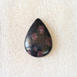 Puede incluir: Una gema pulida en forma de l&aacute;grima. La piedra es predominantemente negra, con varias inclusiones de color granate. La gema se encuentra sobre un fondo blanco texturizado.