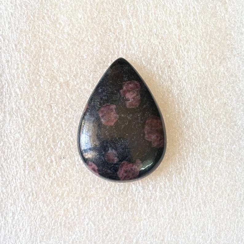 Puede incluir: Una gema pulida en forma de l&aacute;grima. La piedra es predominantemente negra, con varias inclusiones de color granate. La gema se encuentra sobre un fondo blanco texturizado.