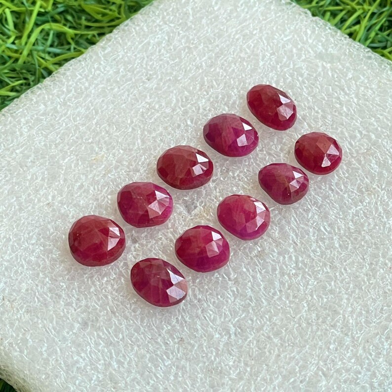 Puede incluir: Una colecci&oacute;n de diez piedras preciosas rojas de forma ovalada y facetadas, dispuestas en un patr&oacute;n escalonado. Cada piedra presenta una superficie pulida con m&uacute;ltiples facetas geom&eacute;tricas que reflejan la luz, mostrando un color carmes&iacute; profundo y uniforme.