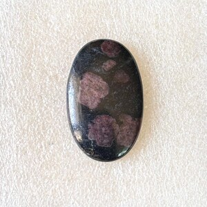 May include: An oval-shaped gemstone with a polished surface. The stone is primarily black with irregular patches of deep pink and purple hues. The gemstone is set against a white, textured background.