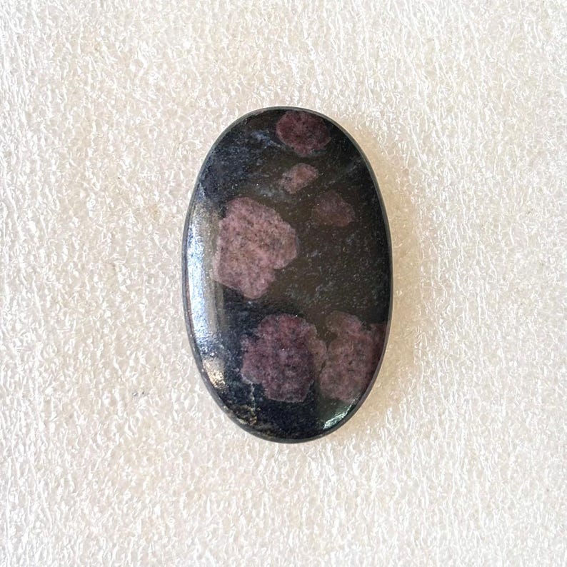 May include: An oval-shaped gemstone with a polished surface. The stone is primarily black with irregular patches of deep pink and purple hues. The gemstone is set against a white, textured background.