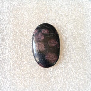 Puede incluir: Gema de forma ovalada con superficie pulida. La piedra es principalmente negra con varias inclusiones circulares de color rosa oscuro. La gema est&aacute; sobre un fondo blanco texturizado.