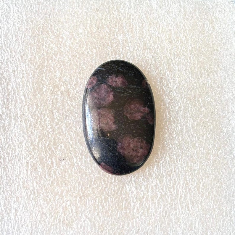 Puede incluir: Gema de forma ovalada con superficie pulida. La piedra es principalmente negra con varias inclusiones circulares de color rosa oscuro. La gema est&aacute; sobre un fondo blanco texturizado.