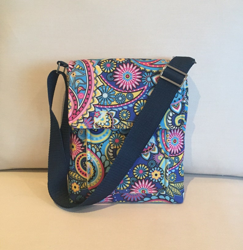 May include: A colorful crossbody bag with a paisley pattern in shades of blue, pink, yellow, and purple. The bag has a flap closure and a navy blue adjustable shoulder strap. The bag is made of fabric.