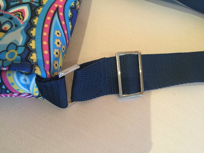 May include: Close-up of a section of a patterned bag with a navy blue strap. The bag features a colorful paisley design with blue, pink, and yellow accents. A silver-colored metal buckle connects the strap to the bag.