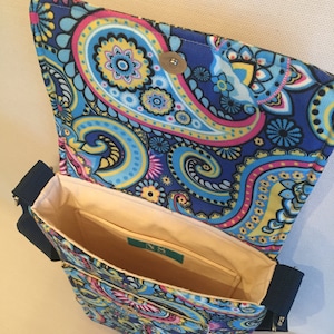 May include: A blue paisley pattern crossbody bag with a flap closure. The bag features a vibrant design with pink, yellow, and blue swirls. The interior is beige with a small pocket and a label. The bag has a navy blue strap and zipper.