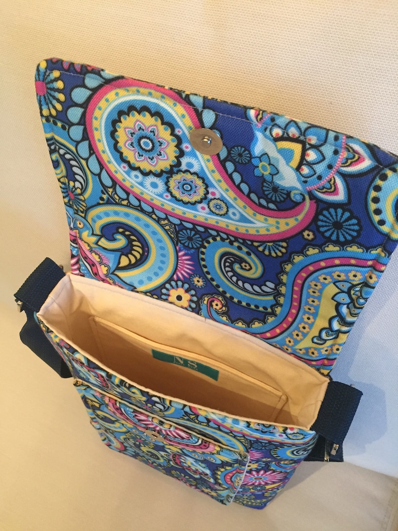 May include: A blue paisley pattern crossbody bag with a flap closure. The bag features a vibrant design with pink, yellow, and blue swirls. The interior is beige with a small pocket and a label. The bag has a navy blue strap and zipper.