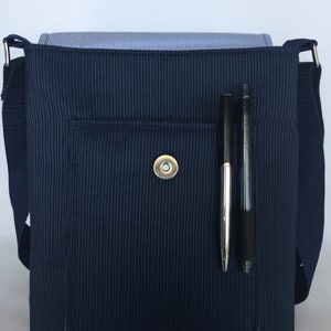 May include: A navy blue messenger bag with a vertical ribbed texture. The bag has a front pocket with a magnetic snap closure. Two pens are in a pen holder on the front. A blue and white striped shirt is inside the bag.