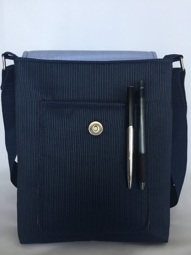 May include: A navy blue messenger bag with a vertical ribbed texture. The bag has a front pocket with a magnetic snap closure. Two pens are in a pen holder on the front. A blue and white striped shirt is inside the bag.