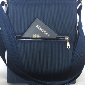 May include: Navy blue messenger bag with a textured, vertical ribbed pattern. The bag features a zippered front pocket with a gold-colored zipper pull. A passport is partially visible in the pocket. The bag has a matching navy blue adjustable shoulder strap.