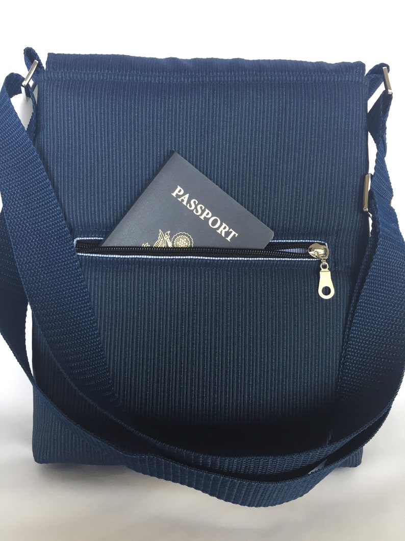 May include: Navy blue messenger bag with a textured, vertical ribbed pattern. The bag features a zippered front pocket with a gold-colored zipper pull. A passport is partially visible in the pocket. The bag has a matching navy blue adjustable shoulder strap.
