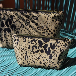 Könnte beinhalten: Zwei Kosmetiktaschen mit Leopardenmuster und goldenen Reißverschlüssen liegen auf einem türkisfarbenen Stuhl. Die Taschen haben einen beigefarbenen Hintergrund mit dunkelblauen Flecken. Die Reißverschlüsse sind goldfarben.