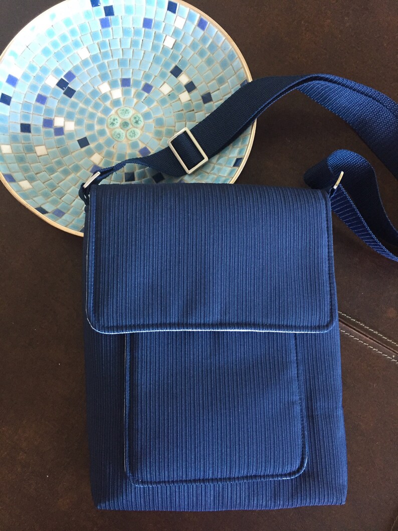 May include: A navy blue crossbody bag with a vertical ribbed texture, a flap closure, and a front pocket. The bag has a matching adjustable strap and silver-colored hardware. A decorative mosaic bowl is in the background.