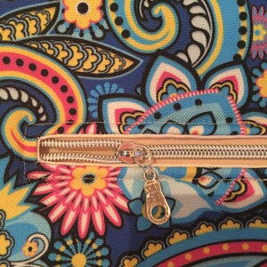 May include: Close-up of a fabric with a vibrant paisley pattern in shades of blue, yellow, pink, and black. A beige zipper with a silver pull is visible across the image, adding a functional element to the design.