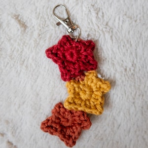 Op de afbeelding: Een sleutelhanger met drie gehaakte sterren in rood, oranje en geel. De sterren zijn bevestigd aan een zilveren metalen sleutelring.
