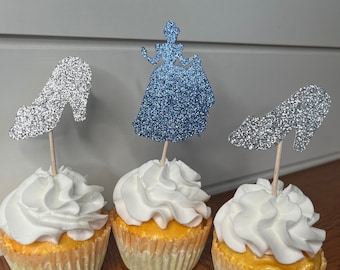 May include: Three cupcakes with white frosting and blue and silver glitter Cinderella themed toppers. The toppers are a silhouette of Cinderella in a blue dress and two silver glass slippers.