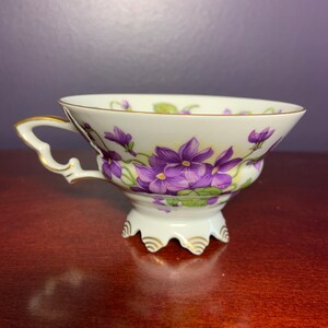 May include: A white teacup with a footed base and a delicate handle. The cup is decorated with a floral pattern of purple violets and green leaves. The rim of the cup is accented with a thin gold band.