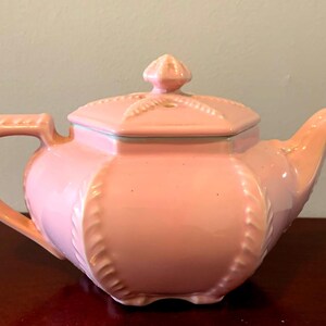 May include: A pink ceramic teapot with a lid and a decorative design. The teapot has a rounded body and a spout on the side.